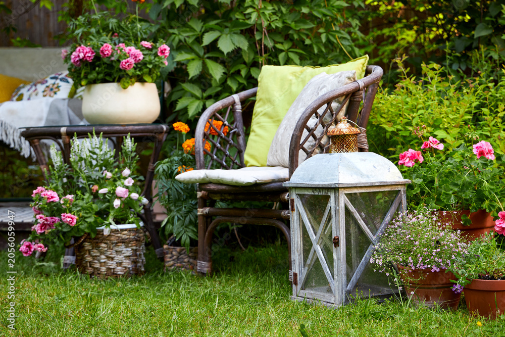 Fototapeta premium Charming outdoor garden seating area featuring a wicker chair surrounded by colorful blooming flowers, decorative lanterns, woven baskets, and lush green foliage. This peaceful backyard