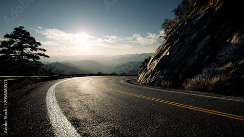 A scenic, winding mountain highway with double yellow lines, hugging a steep rocky cliff on the right, overlooking misty hills under a bright afternoon sun