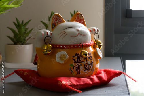 Cute ceramic cat sits on red fabric. Ears and paws feature small golden bells. Chinese characters decorate its belly. Sitting near a potted plant, it looks serene