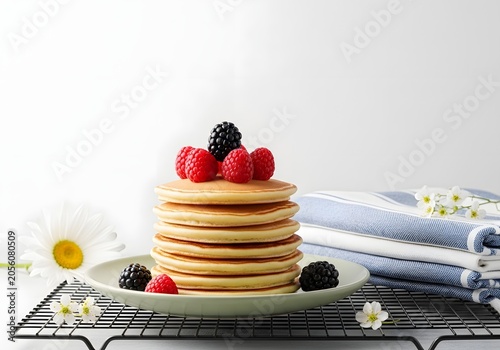 A bountiful stack of fluffy, golden pancakes adorned with fresh raspberries and blackberries, artfully presented on a ceramic plate beside delicate white daisies and folded checkered linens