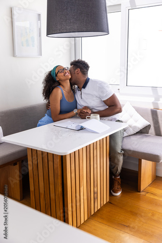 Diverse couple kissing and holding hands at home dining nook with coffee mug notebook