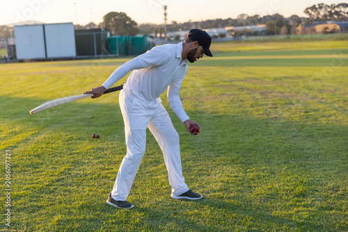 Cricket bat is leaning while red balls are resting on mown cricket field with long shadows