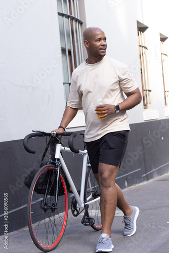 African American man in sportswear walking along masonry wall holding road bike and orange juice