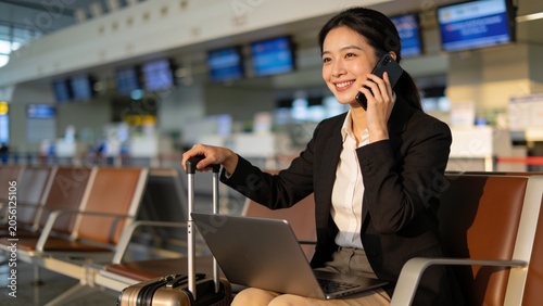 Asian female sitting at airport before trip having telephonic conversation with client and smiling. 
