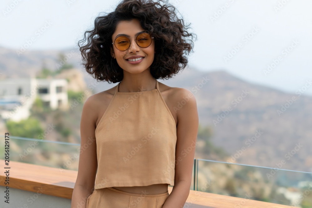 Fototapeta premium Woman in tan halter crop top and high waisted trousers on balcony with hillside view for lifestyle portrait background and vacation banner