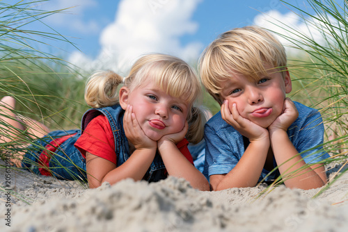 two children lying on sandy ground with green dune grass around them under a blue sky with white clouds. They sticking their tongs out.