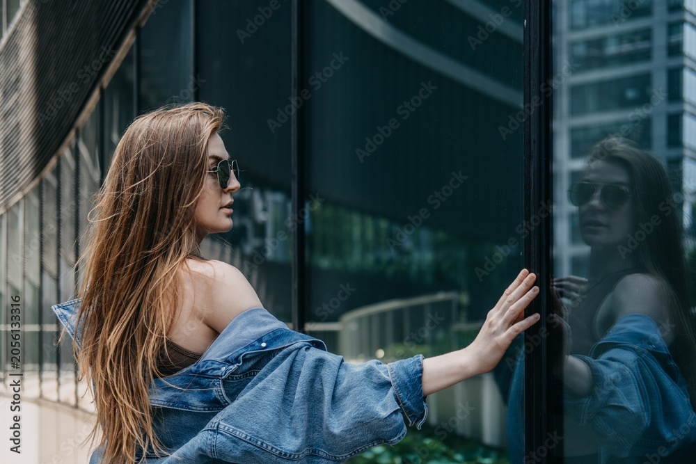 Fototapeta premium Woman wearing sunglasses looks at her reflection in glass facade of modern office building. self reflection concept, introspective business professional, identity in modern city.