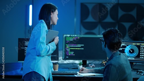 Two Asian software engineers collaborate in a dark modern office. Male programmer points at code on the computer screen, explaining data to his female IT colleague holding a tablet. Teamwork concept.