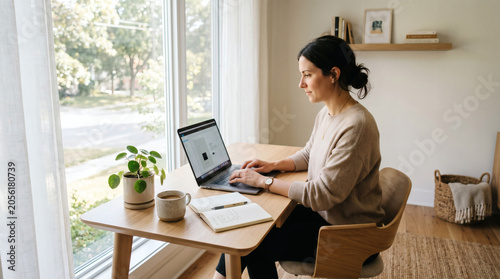Smiling young woman using laptop at bright home office window in modern apartment
