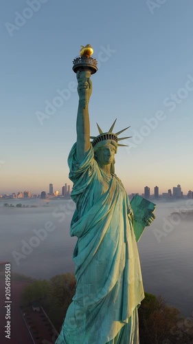 Slow vertical drone pan of Statue of Liberty at sunrise, New York. Low sea mist, clear sky. Manhattan skyline is visible. Copper patina illuminated monument.