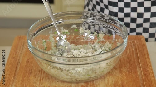 A chef mixing minced fish with egg and chopped fresh herbs in a glass bowl to prepare a mixture for fish patties