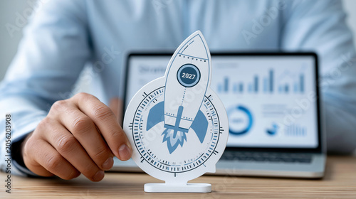 Rocket launch concept with 2027 year, business growth and success plan, hand holding paper rocket in front of laptop screen with charts