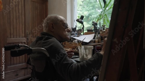 Senior artist on a wheelchair paints an oil painting on canvas in a studio