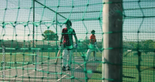 Adult indian batter gripping bat in cage as teenage trainee tossing baseballs, swing practice