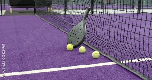 Opening shot framing black paddle leaning on net with three balls on purple court next to bench