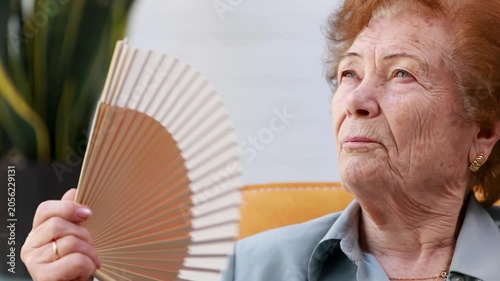 portrait of a senior woman fanning herself with a fan while seated indoors; concept of summer heat wave and elderly people