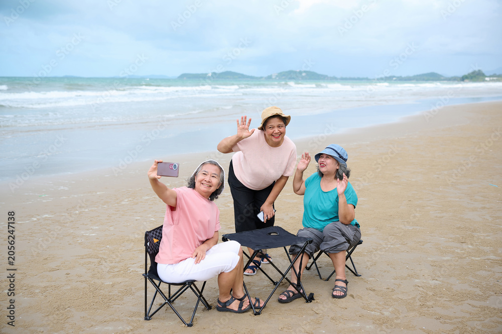 Obraz premium Elderly women happily enjoying a day at the beach, taking selfies and enjoying each other's company. They are sitting on foldable chairs at a small table on the sand.