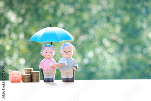 Senior couple figures under blue umbrella with stacks of coins on green background. Concept for retirement insurance, financial protection, elderly wealth management and pension security.