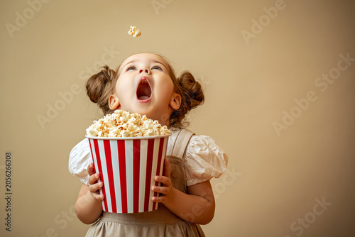 Playful young girl catching popcorn mid air with mouth open holding a red and white striped bucket on a warm beige background.