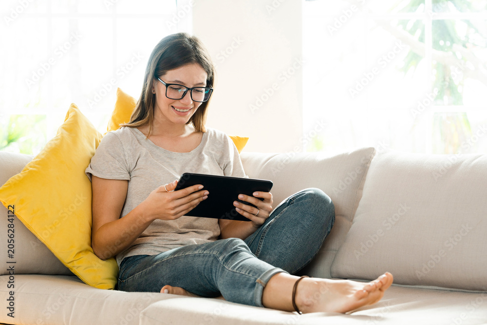 © Westend61 - Smiling brunette woman with glasses relaxing on couch and looking at her tablet