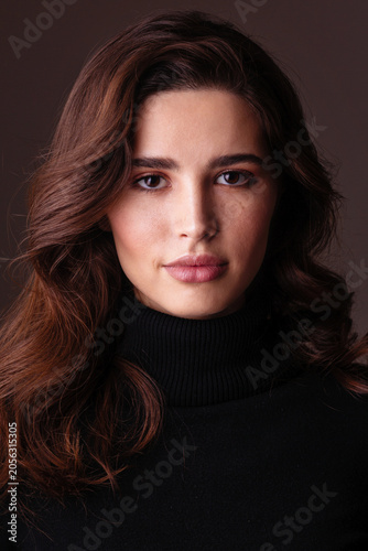 A young woman with long wavy brunette hair wears a black turtleneck sweater and looks confidently at the camera in a studio portrait. Soft makeup and a calm expression create a timeless beauty image.