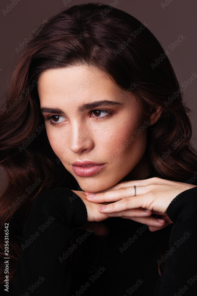 Fototapeta premium A thoughtful woman with brunette hair poses in a beauty portrait wearing a black sweater. Soft studio lighting highlights natural makeup, smooth skin, expressive eyes against a warm background.