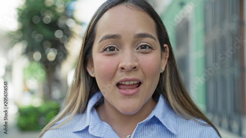Fotografija Young hispanic woman mouth open smiling with visible teeth and rosy cheeks on a city street closeup, wearing blue striped shirt and necklace; joy warmth