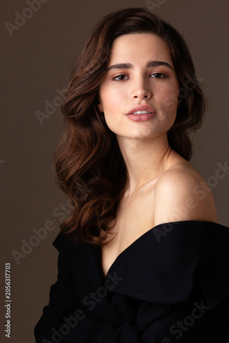 A young brunette woman with wavy hair poses in an elegant black off-shoulder dress against a neutral studio background. Her natural beauty, confident expression, and refined style create a beauty 