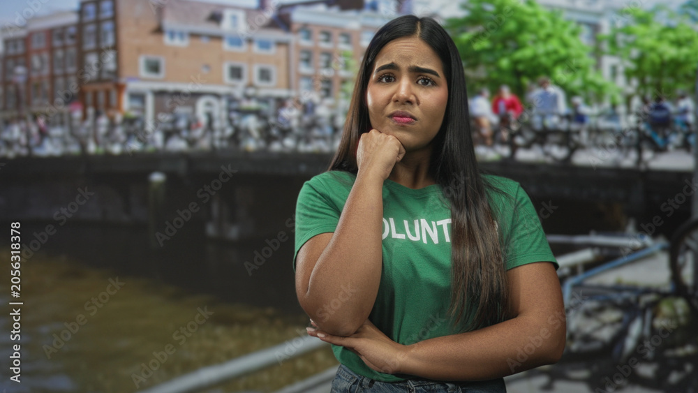 Fototapeta premium Young woman volunteer with hand on chin and crossed arms on a street by a canal and bicycles; concern reflection.
