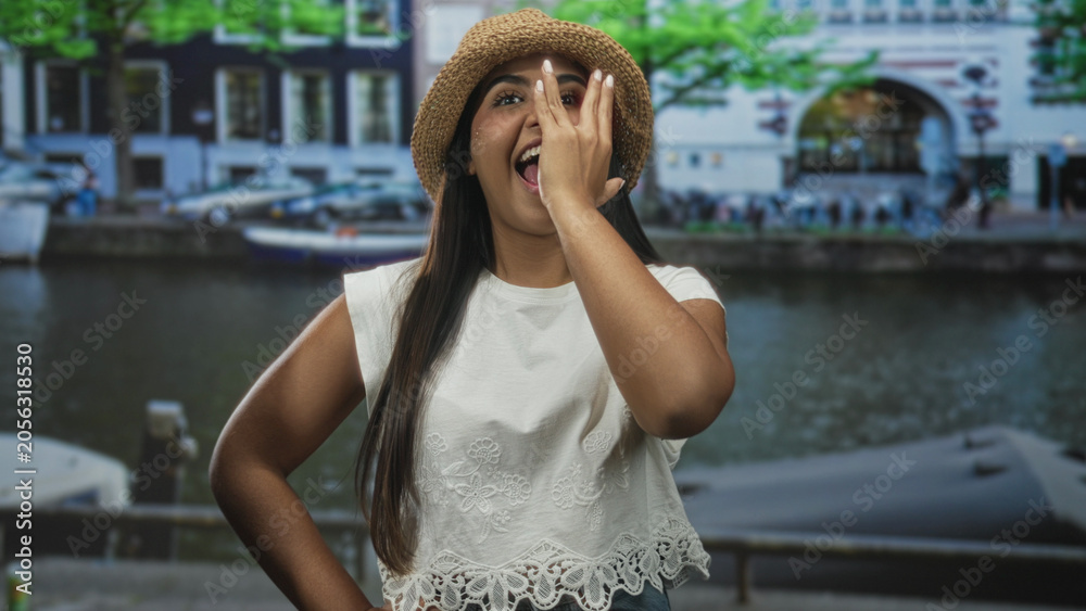 Fototapeta premium Woman covering one eye with hand while smiling in front of a canal building, wearing straw hat and white lace top; playful joy.