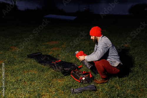 Photos Young bearded man packing sleeping bag or checking equipment before pitching tourist tent on lake shore at dark twilight night