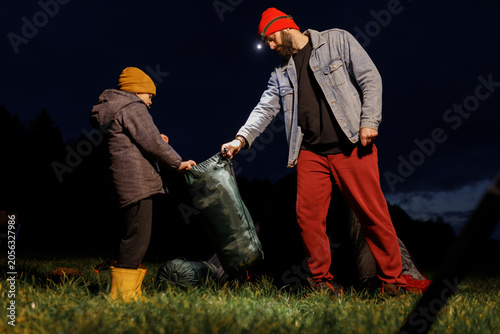 Photos Young bearded father with headlamp flashlight packing sleeping bag or camping gear with little son on lake shore at dark night
