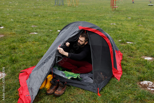 Quadro su tela Young bearded man opening zipper of tourist camping tent entrance on green grass field in morning