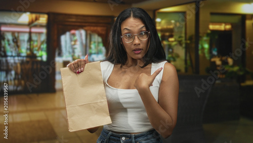 Fotografía Woman holding brown paper bag, waving open hand and pointing to chest while smiling in restaurant dining area with tables, chairs and plants; surprise delivery