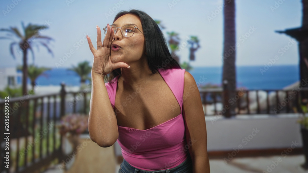 Fototapeta premium Woman cups hands to shout on building balcony, mouth open, wearing pink top, glasses and denim jeans; playful excitement.