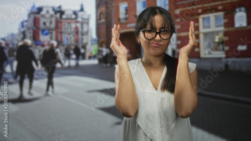 Φωτογραφία Young latina woman with hands covering eyes and pursed lips on busy street; embarrassment hesitation doubt