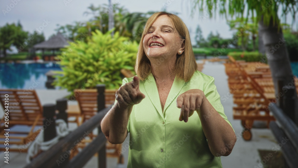 Fototapeta premium Woman pointing fingers and smiling toward camera on a pool deck near lounge chairs and railing in a building; joyful connection.