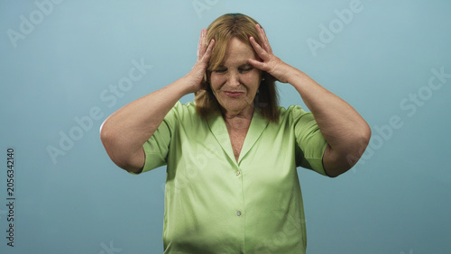 Fotografie Middle age woman holding head with hands, palms on forehead, wearing green shirt, studio backdrop; anxiety worry