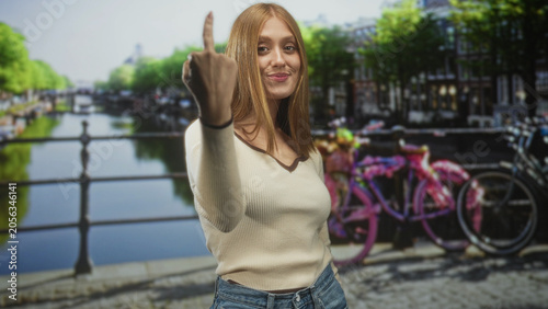 Foto Woman shows middle finger on a canal bridge beside pink bicycles, wearing cream sweater and jeans; defiance attitude