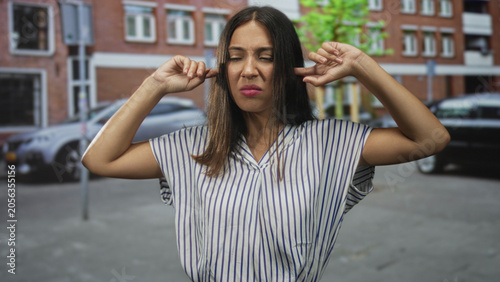 Принти на полотні Young woman plugging ears with fingers on city street in front of brick buildings and parked cars; noise annoyance