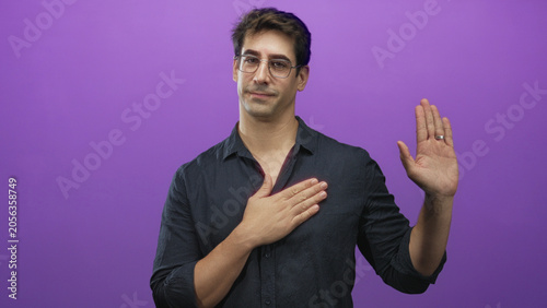Fényképezés Man wearing glasses and dark shirt, hand placed over chest and other hand raised in oath gesture in studio purple backdrop with wedding ring visible; sincerity