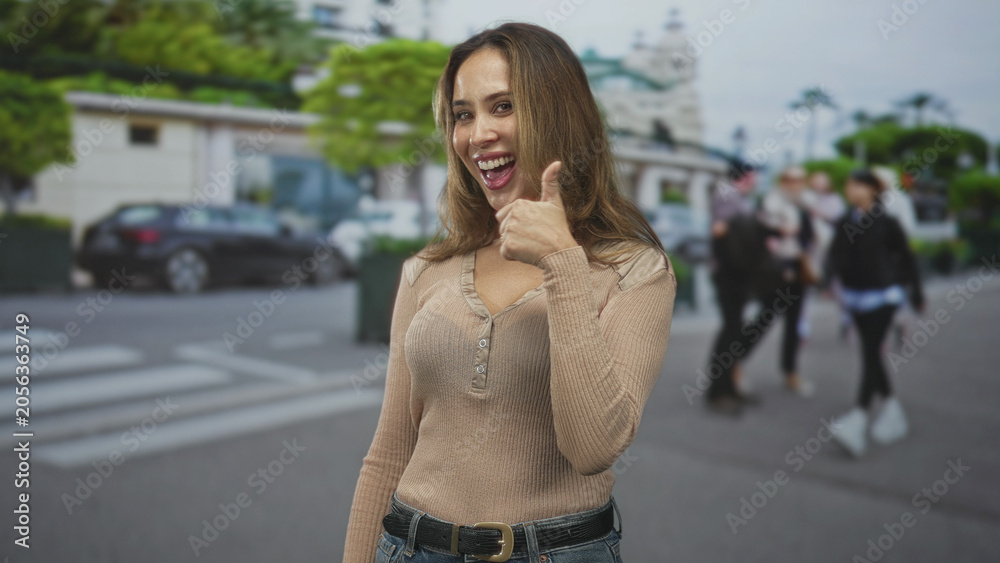 Fototapeta premium Woman pointing finger at street crosswalk while smiling, visible hand, beige sweater and belt with parked car and pedestrians nearby; happy approval.