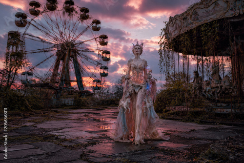 A woman in an elaborate, whimsical dress stands amidst an overgrown amusement park at sunset. Eerie beauty and nostalgia.
