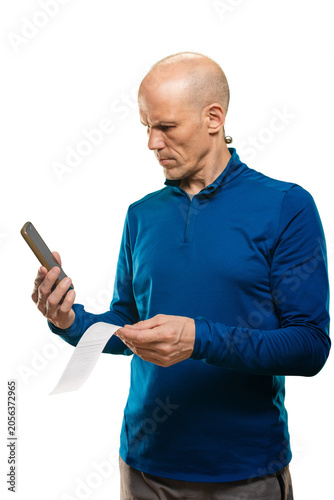 Man comparing receipt against smartphone, frontal view of shopper reviewing totals and prices, blue top visible, scanning list of items and quantities, moment of uncertainty about