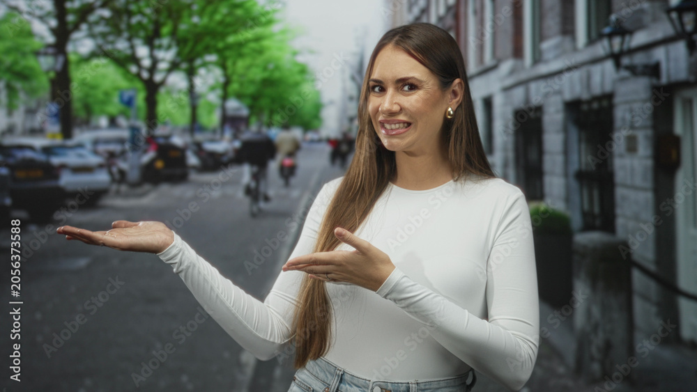 Fototapeta premium Woman presenting palm and gesturing with other hand on street with buildings and parked cars; bemusement uncertainty.