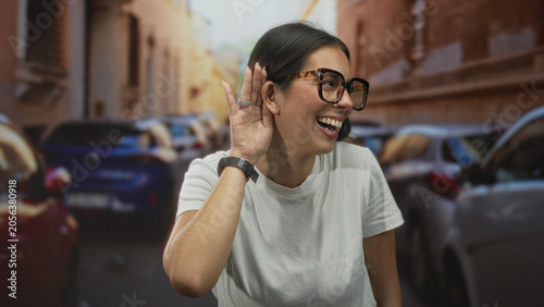 Εκτύπωση καμβά Woman cupping hand to ear on narrow street beside parked cars, wearing glasses and white t shirt; joy