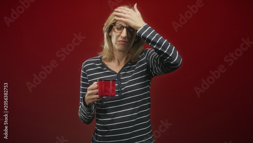 Obraz na plátně Woman holding red coffee mug coughs and covers mouth while grimacing in studio; discomfort reaction