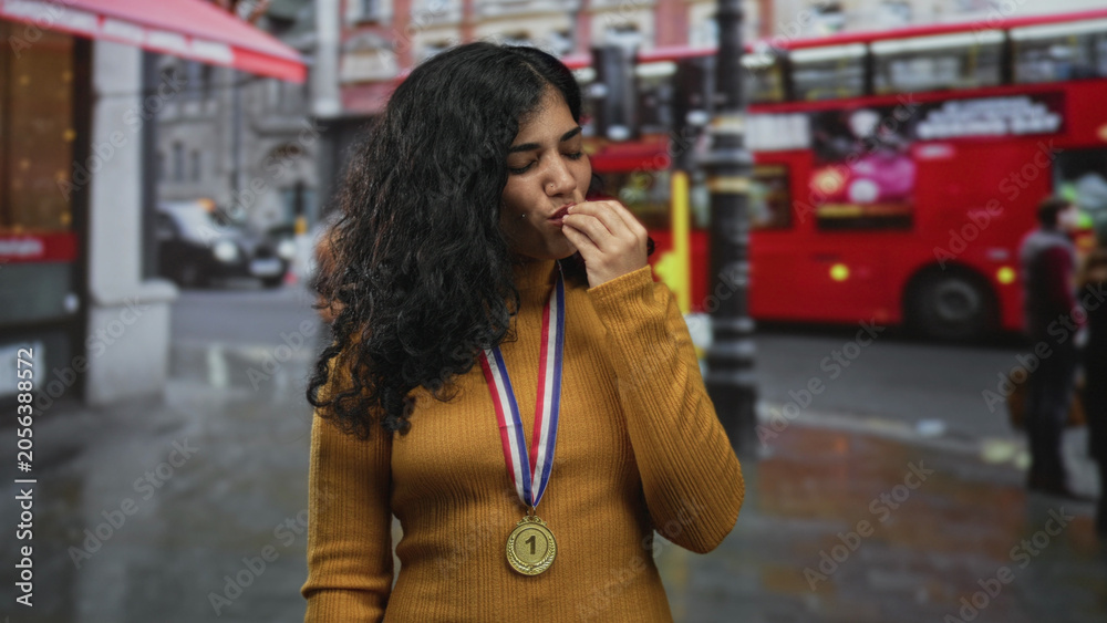 Fototapeta premium Woman wearing first place medal on rainy street in front of red doubledecker bus, hand raised near mouth with eyes closed; victory celebration pride.