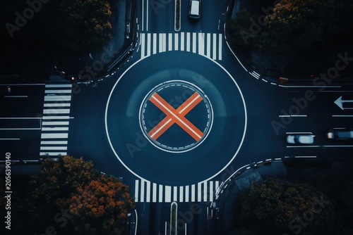 Overhead view of a city roundabout with cars, crosswalks and a bold orange x in the center at night.