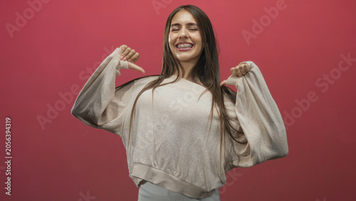 Foto Teen girl with braces and long hair smiling, thumbs pointing to chest while wearing a beige sweater against a pink backdrop in studio; playful confidence